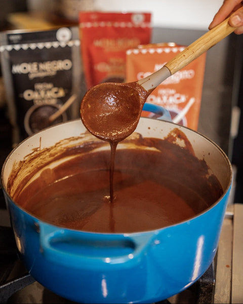 A close-up of a bright blue pot filled with melted dark chocolate, with a wooden spoon lifting some of the chocolate to show its smooth, glossy texture. In the background, out-of-focus packages of chocolate and cocoa powder are visible.